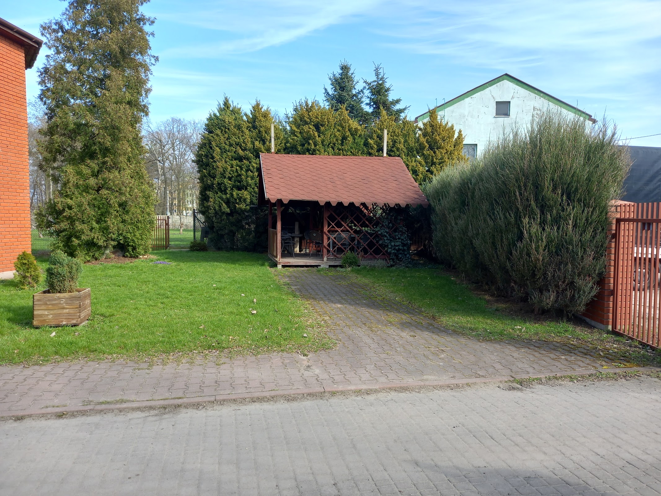 Widok na drewnianą altanę ogrodową z czerwonym dachem i białym budynkiem gospodarczym w tle, brukowa ścieżka, zielone tuje